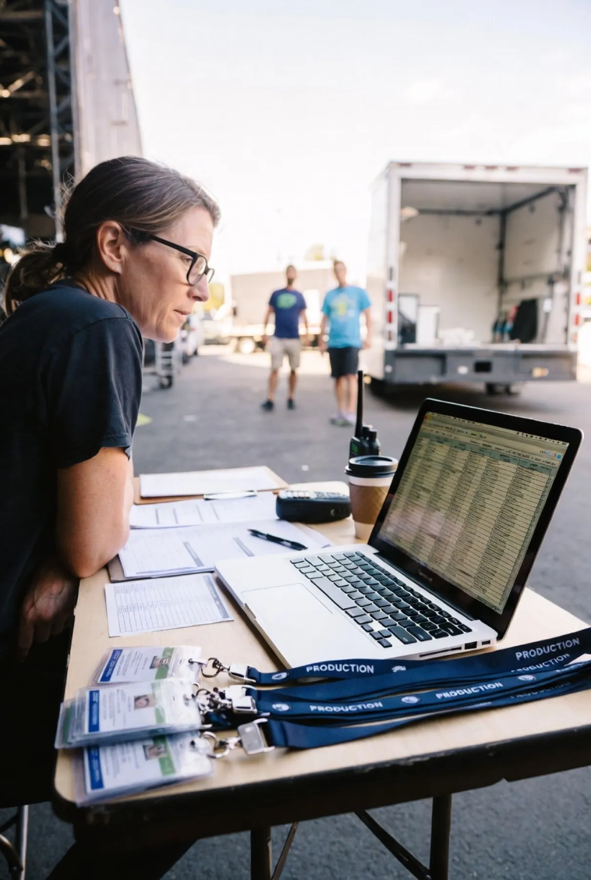 A crew member checks a call sheet and manifest on a laptop at a production table, truck and crew behind her.